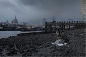 Atmospheric photo of artist Liz Crow sitting on the mudbanks of the Thames river