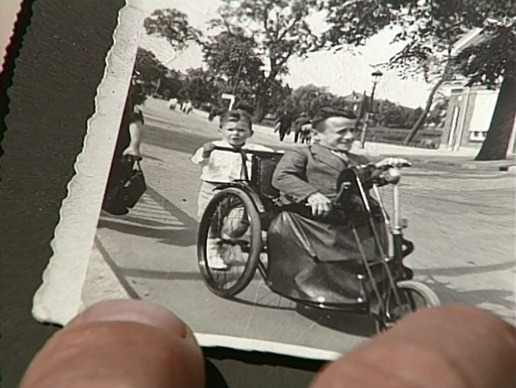 A black and white photograph of a young white male in an old style three wheeled motorised wheelchair is being held up to the camera. 